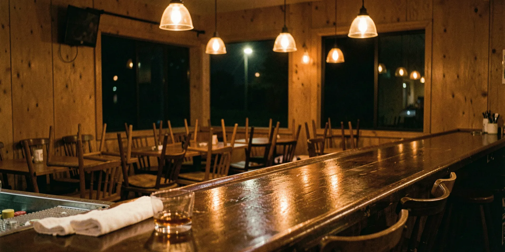 A neighborhood bar after closing: chairs upturned on the tables, a whiskey glass and a folded bar towel on the empty bar under warm pendant lights.